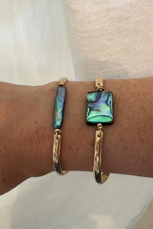 A stack of two gold bangle bracelets with blue pearl and abalone pendants on a person's wrist.